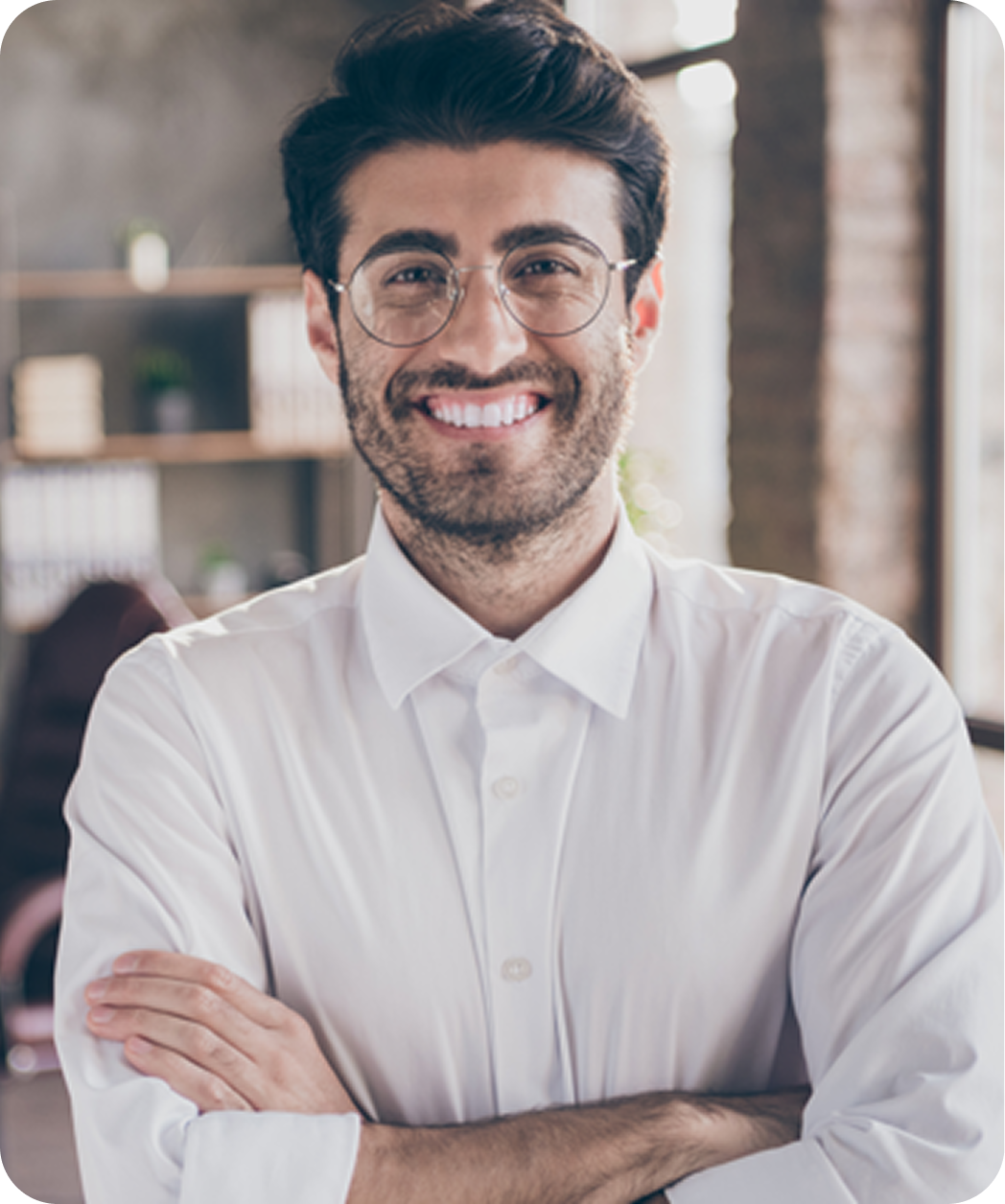 Um homem jovem e sorridente, com cabelo escuro e óculos, está de braços cruzados, vestindo uma camisa branca. Ele está em um ambiente iluminado, com uma decoração de estilo moderno ao fundo.