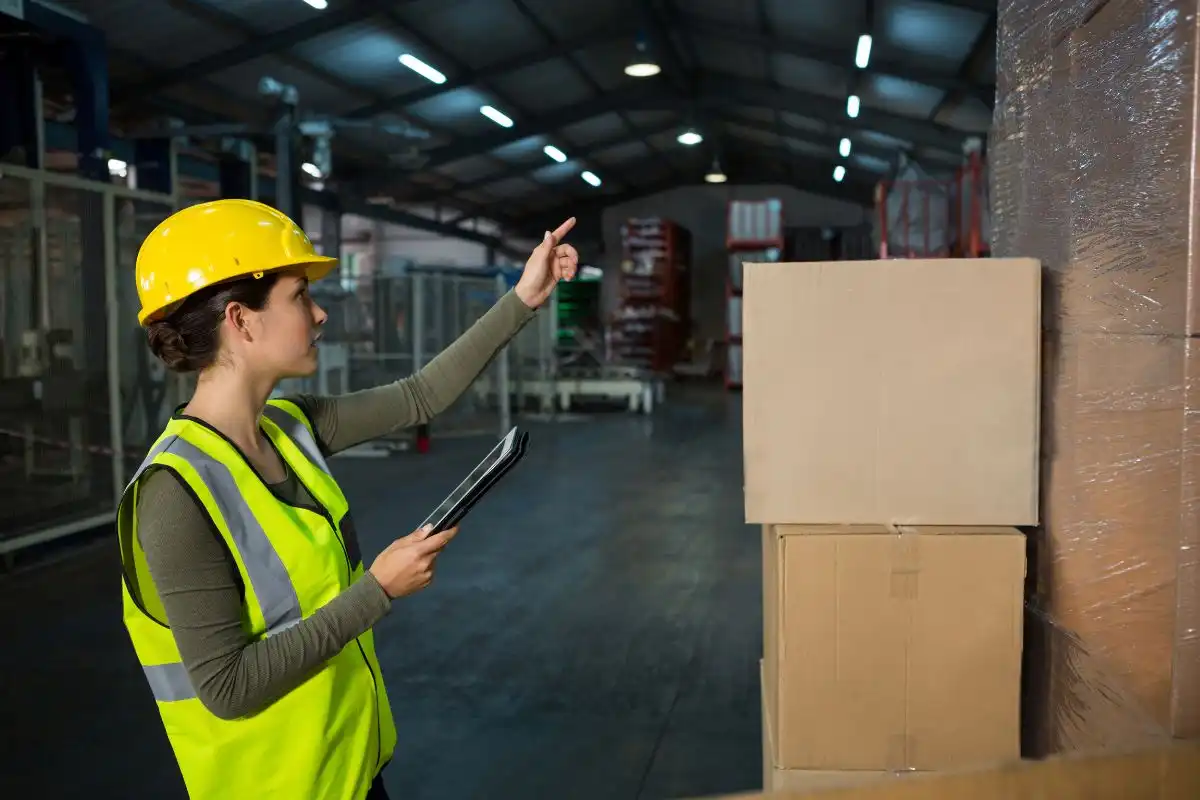 Profissional de logística organizando a movimentação de carga interna, verificando estoque em armazém com caixas de papelão, usando capacete amarelo e vestimenta refletora, no ambiente industrial.